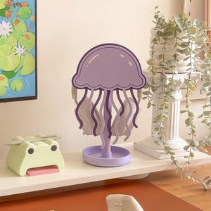 May include: A purple jellyfish-shaped desk organizer with wavy tentacles and a round base. A light green frog-shaped desk organizer with a pink notepad is to the left. A white keyboard and mouse are in the foreground.