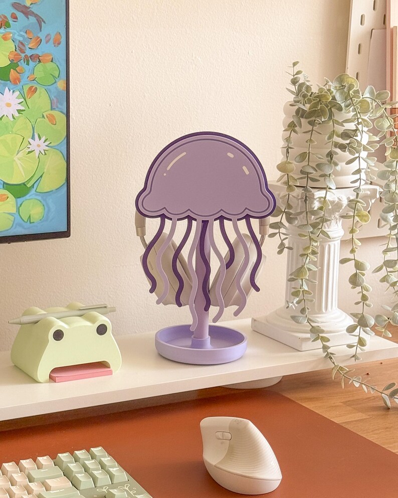 May include: A purple jellyfish-shaped desk organizer with wavy tentacles and a round base. A light green frog-shaped desk organizer with a pink notepad is to the left. A white keyboard and mouse are in the foreground.