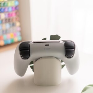 May include: A white and black video game controller with the word "SONY" printed on it, resting on a white stand. The controller has black thumbsticks and buttons. The stand is cylindrical and sits on a white surface.
