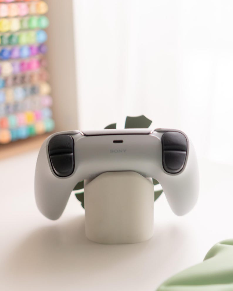 May include: A white and black video game controller with the word "SONY" printed on it, resting on a white stand. The controller has black thumbsticks and buttons. The stand is cylindrical and sits on a white surface.