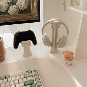 May include: A white desk setup featuring a black game controller and white headphones on decorative stands. A keyboard with white and green keys sits in the foreground. A small teddy bear figurine is also present.
