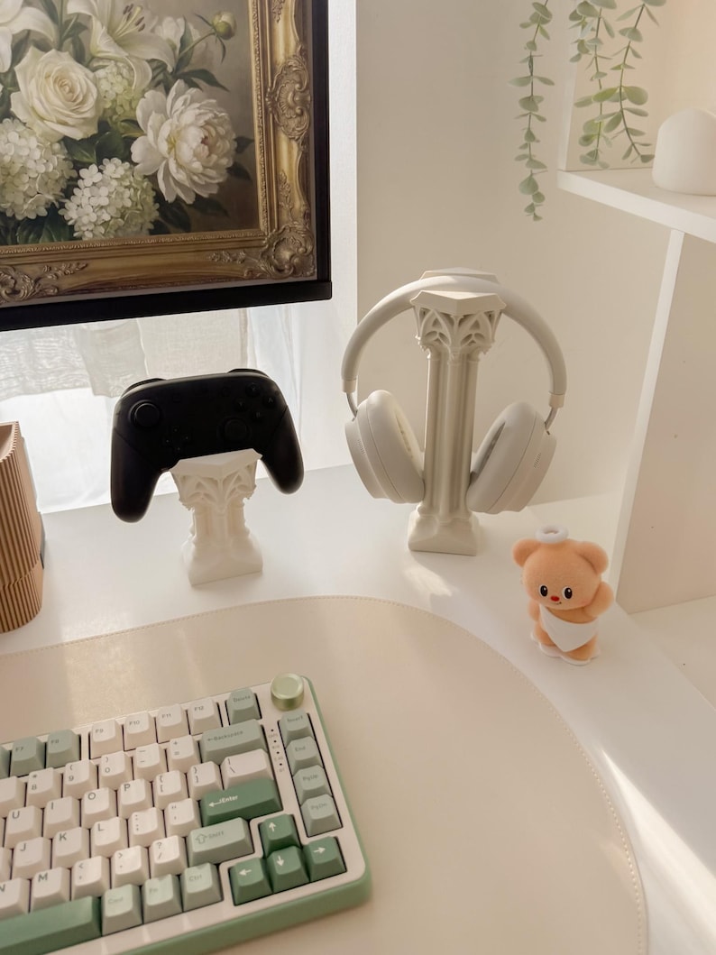 May include: A white desk setup featuring a black game controller and white headphones on decorative stands. A keyboard with white and green keys sits in the foreground. A small teddy bear figurine is also present.