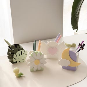 May include: A collection of decorative pen holders in various shapes, including a monstera leaf, daisy, angel wings with a heart, and a moon with a cloud. The pen holders are in pastel colors and are arranged on a light-colored surface.