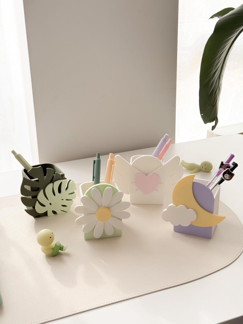 May include: A collection of decorative pen holders in various shapes, including a monstera leaf, daisy, angel wings with a heart, and a moon with a cloud. The pen holders are in pastel colors and are arranged on a light-colored surface.