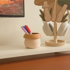 May include: A light pink and tan ceramic pen holder with three colorful markers inside. A mushroom-shaped headphone stand holds white headphones. The items are on a white shelf against a neutral wall.