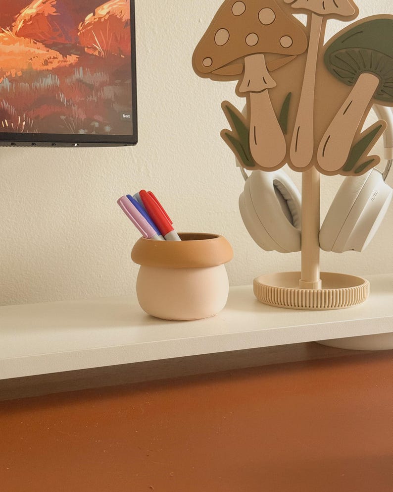 May include: A light pink and tan ceramic pen holder with three colorful markers inside. A mushroom-shaped headphone stand holds white headphones. The items are on a white shelf against a neutral wall.