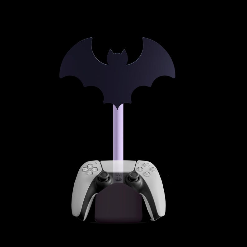 K&ouml;nnte beinhalten: Ein schwarz-wei&szlig;er Gaming-Controller ruht auf einem lila St&auml;nder mit einer Fledermaus-Silhouette. Der St&auml;nder ist so konzipiert, dass er den Controller h&auml;lt, mit einem dunkelvioletten Sockel und einem hellvioletten Stiel.