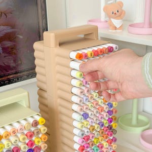 May include: A beige marker organizer filled with colorful markers. The organizer has a handle and a ribbed design. A hand is reaching for a marker. The markers are arranged by color, with a variety of shades visible.