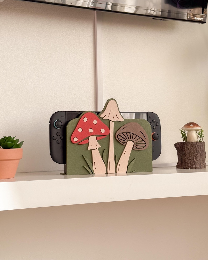 Pu&ograve; includere: Un supporto per console di gioco in legno con un design a forma di fungo. Il supporto &egrave; dipinto di verde con funghi tridimensionali rossi, marroni e beige. Dietro il supporto &egrave; posizionata una console di gioco nera.
