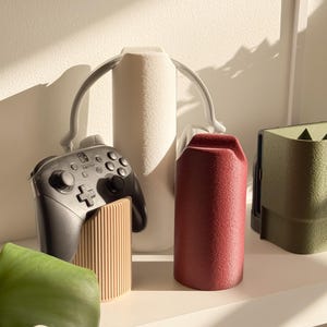 Może przedstawiać: Zestaw akcesoriów do gier, w tym czarny kontroler Nintendo Switch Pro, beżowa podstawka pod kontroler oraz cylindryczne pojemniki w kolorze białym, czerwonym i zielonym. Przedmioty są ustawione na białej półce.