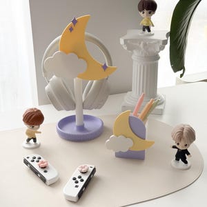 Puede incluir: Un juego de organizadores de escritorio con un diseño de luna creciente y nube amarillas. Incluye un soporte para auriculares, un portalápices y figuritas. El conjunto es de colores pastel e incluye dos mandos de juego blancos con detalles de flores rosas.