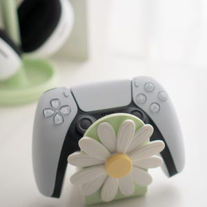 May include: A white and black PlayStation 5 controller rests on a light green stand with a white daisy flower design. The controller has gray accents and a black center.