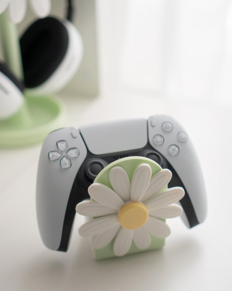 May include: A white and black PlayStation 5 controller rests on a light green stand with a white daisy flower design. The controller has gray accents and a black center.
