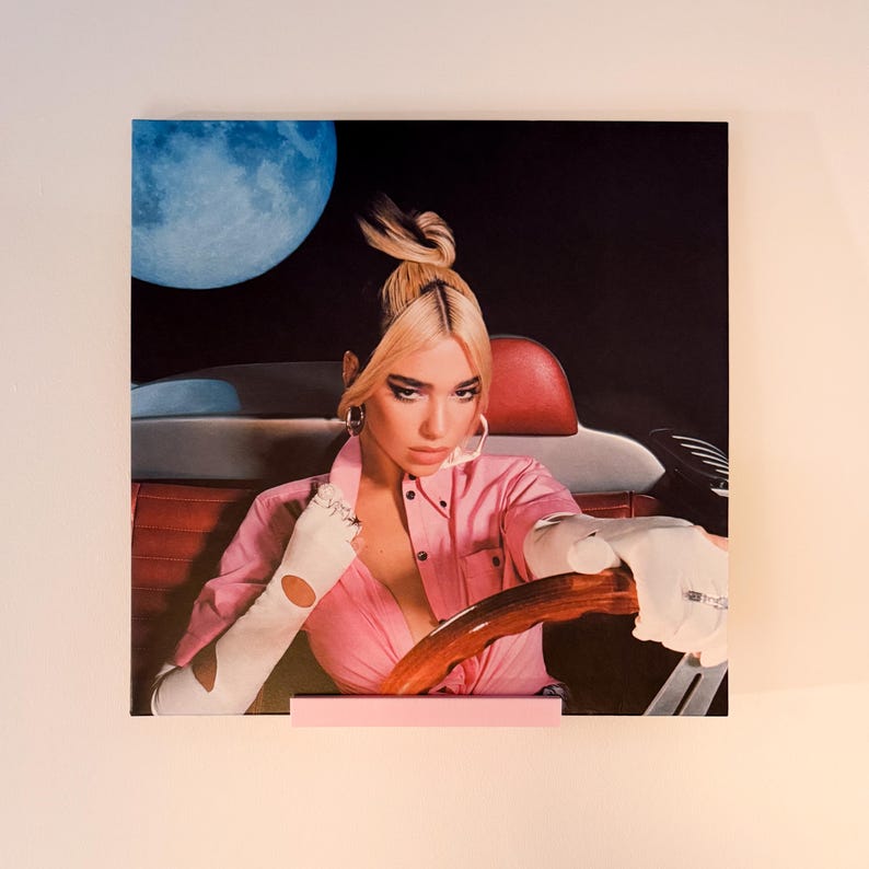 May include: A square print of a woman in a pink shirt and white gloves, driving a car with a wooden steering wheel. A large blue moon is in the background. The print is displayed on a pink stand.
