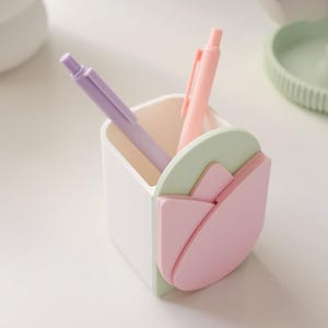 May include: A white pen holder with a tulip design in pink and green, holding a purple and a pink pen. The pen holder is square-shaped and sits on a white surface. The pens are standard size.
