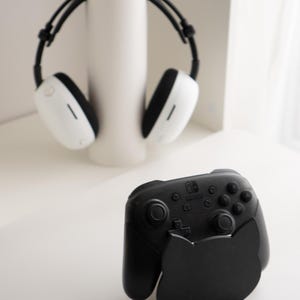 May include: A black Nintendo Switch Pro controller with a cat-shaped stand. The controller has buttons and joysticks. In the background, a pair of white and black headphones rest on a white cylindrical stand.