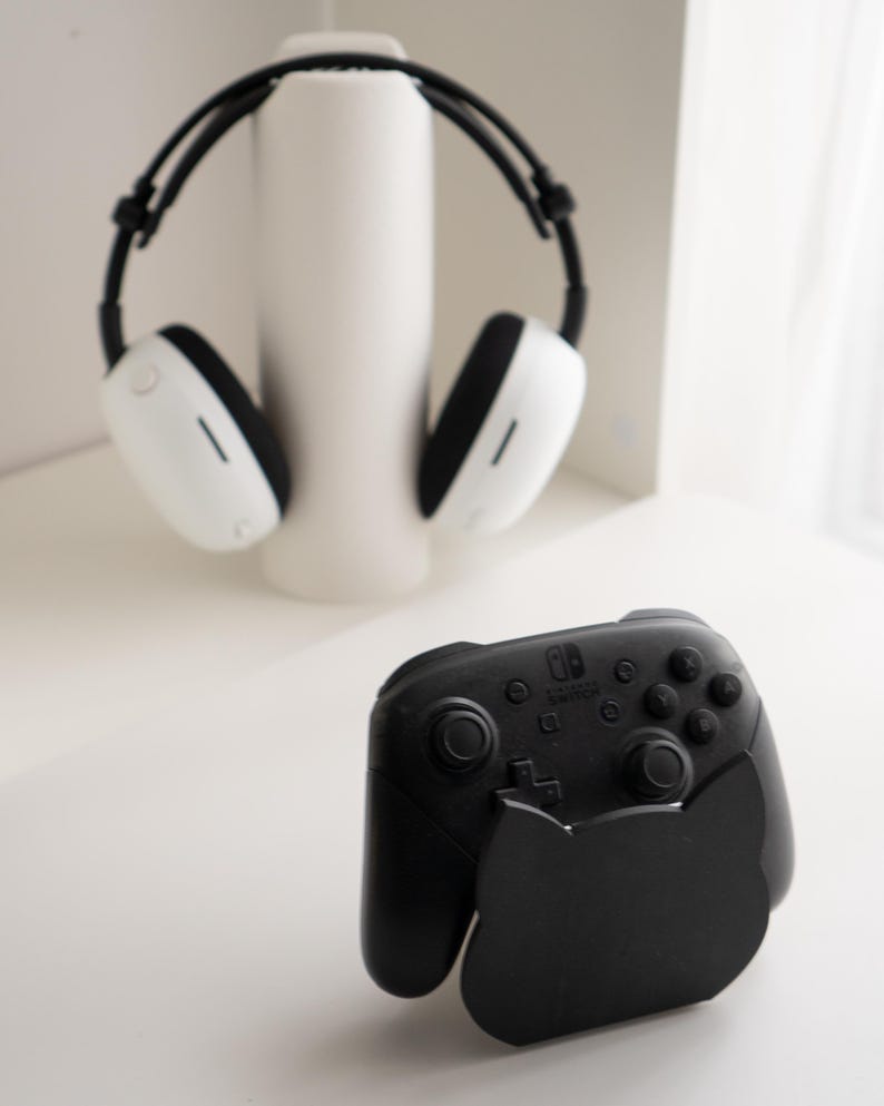 May include: A black Nintendo Switch Pro controller with a cat-shaped stand. The controller has buttons and joysticks. In the background, a pair of white and black headphones rest on a white cylindrical stand.