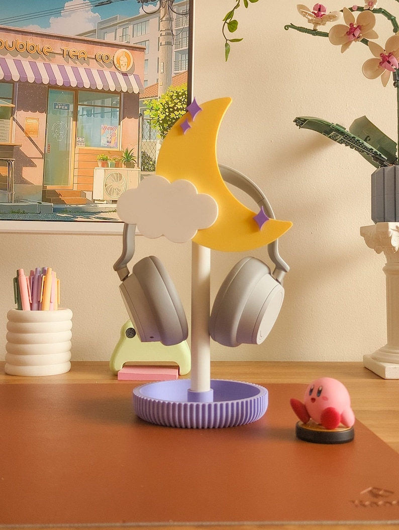 May include: A purple and white headphone stand shaped like a crescent moon with a cloud. The stand has a purple base and is holding two gray headphones.