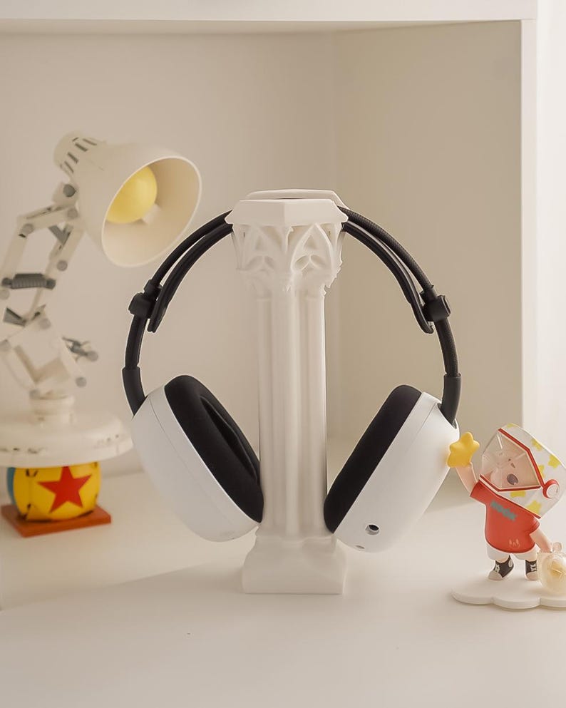 May include: White and black headphones resting on a white decorative column. The headphones have a modern design. A small desk lamp and a figurine are in the background.