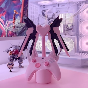 May include: A pink gaming controller stand with a heart-shaped base and black wings. The stand is designed to hold a gaming controller and headphones.