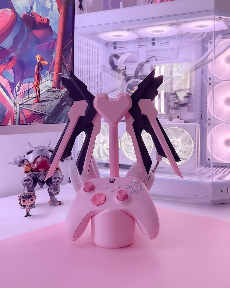May include: A pink gaming controller stand with a heart-shaped base and black wings. The stand is designed to hold a gaming controller and headphones.