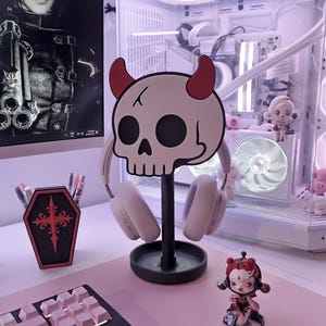 May include: A black headphone stand shaped like a skull with red horns, holding white headphones. A black and red coffin-shaped pen holder and a figurine are on a pink desk. A computer and a poster are in the background.