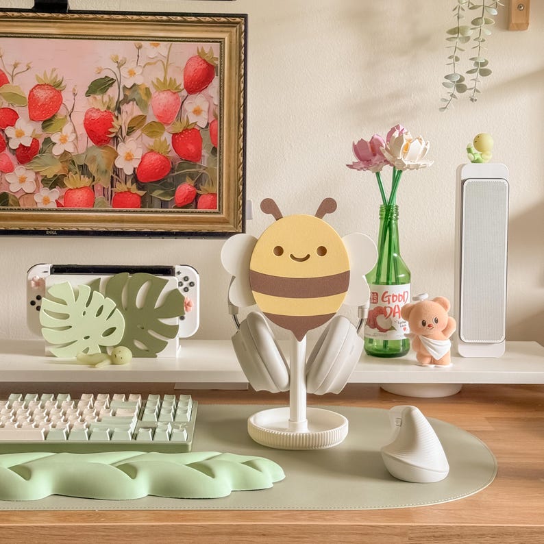 May include: A whimsical desk setup featuring a bee-shaped headphone stand with headphones. A framed strawberry painting hangs above a white shelf holding a gaming console and decorative leaves. A keyboard, mouse, and other decor complete the scene.