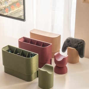 May include: A collection of modern desk organizers in various colors. The set includes a green organizer with compartments, a burgundy organizer, a tan organizer, and a red and green stand. A black game controller rests on a wooden stand.