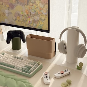 May include: A modern desk setup featuring a computer monitor displaying a video game scene. The desk includes a keyboard, controllers, headphones, and decorative items in neutral tones. A green leaf-shaped desk mat adds a pop of color.