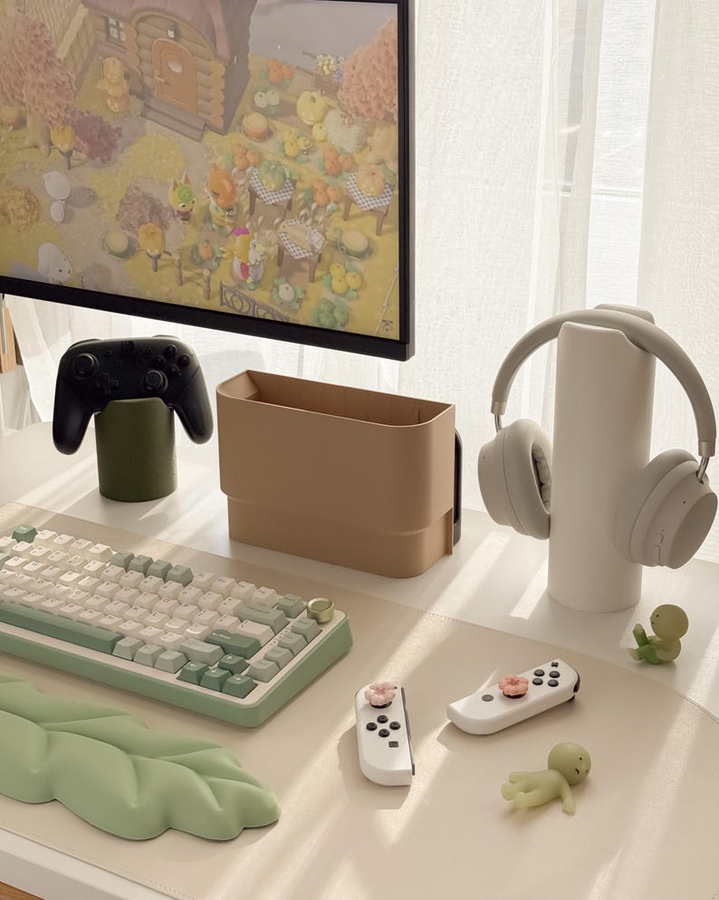 May include: A modern desk setup featuring a computer monitor displaying a video game scene. The desk includes a keyboard, controllers, headphones, and decorative items in neutral tones. A green leaf-shaped desk mat adds a pop of color.
