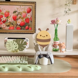 May include: A decorative scene featuring a framed strawberry painting, a bee-shaped controller holder, a white and black game controller, and a green keyboard. A green bottle holds pink flowers, and a small bear figurine is also present.