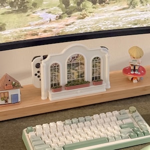 May include: A white plastic gaming console dock shaped like an arched window with three panes, featuring small red flower boxes at the base. The dock is designed to hold a handheld gaming device and is displayed alongside a small house figurine and a mushroom.