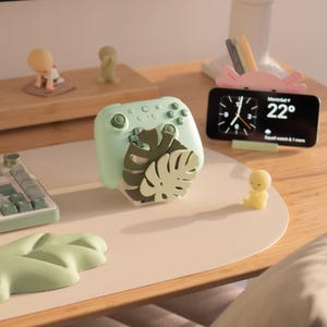 May include: A light green video game controller stand featuring a layered monstera leaf design in dark and light green. The stand holds a matching light green controller and is positioned on a desk with a keyboard, a phone stand, and small figurines.