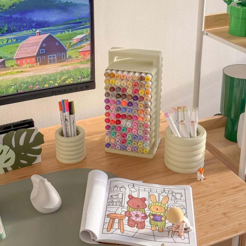 May include: A desk setup featuring a large marker organizer filled with colorful markers. Two small, ribbed, light green containers hold additional markers. A coloring book with cartoon characters is open on the desk, along with a white ergonomic mouse.
