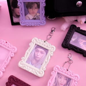 May include: A collection of decorative photo frame keychains in various colors, including lavender, white, pink, and burgundy. Each frame holds a photograph and is attached to a silver keychain. The frames have ornate, curved edges.
