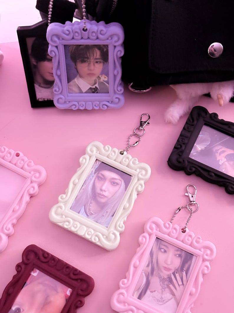May include: A collection of decorative photo frame keychains in various colors, including lavender, white, pink, and burgundy. Each frame holds a photograph and is attached to a silver keychain. The frames have ornate, curved edges.
