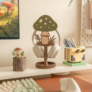 Pu&ograve; includere: Un'impostazione da scrivania stravagante con un supporto per cuffie a forma di fungo nei toni del verde e del marrone. Una piccola statuina a forma di fungo &egrave; appoggiata su una base in finto legno. Sono visibili anche un portapenne ispirato a Minecraft e un mouse ergonomico bianco.