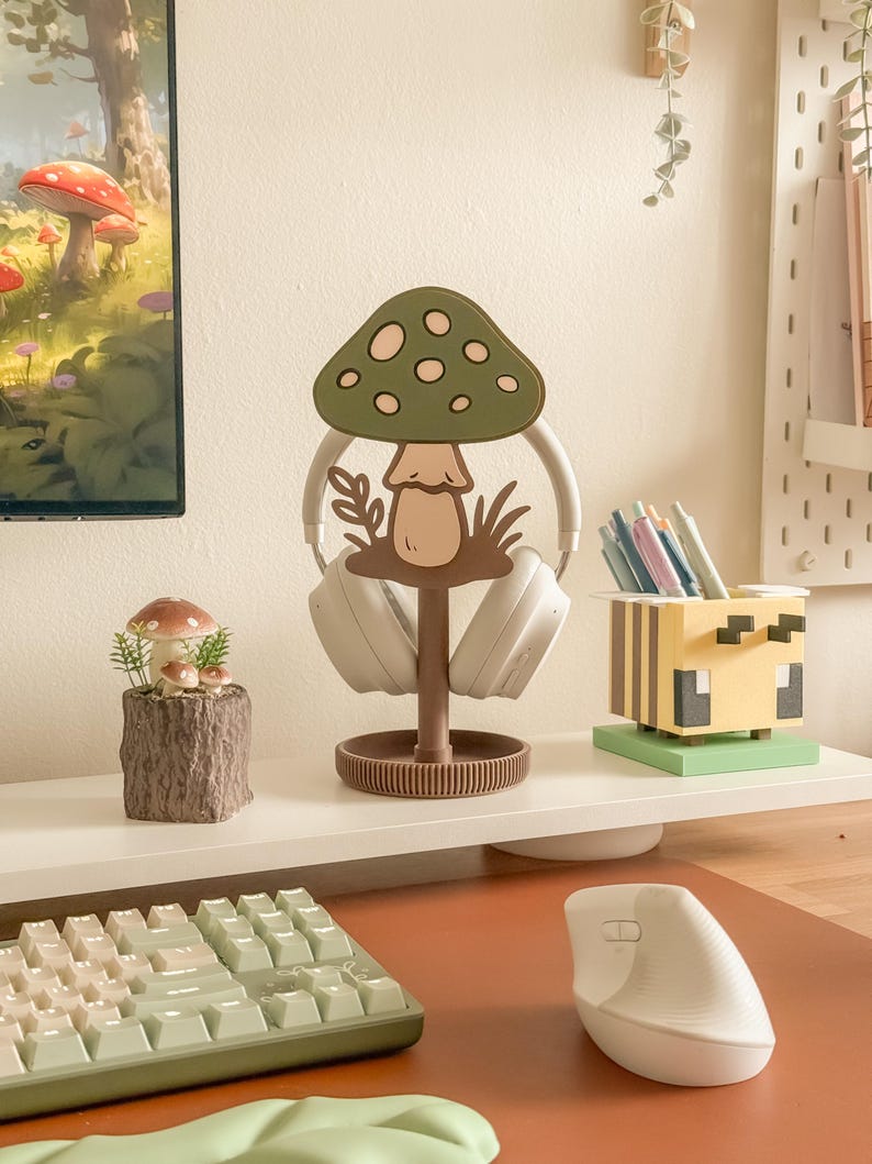 Pu&ograve; includere: Un'impostazione da scrivania stravagante con un supporto per cuffie a forma di fungo nei toni del verde e del marrone. Una piccola statuina a forma di fungo &egrave; appoggiata su una base in finto legno. Sono visibili anche un portapenne ispirato a Minecraft e un mouse ergonomico bianco.
