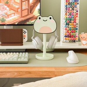 May include: A light green frog-shaped headphone stand holding white headphones. A white keyboard, mouse, and pen holder are on a desk. A white marker holder with colorful markers is in the background. A computer monitor displays a cartoon scene.