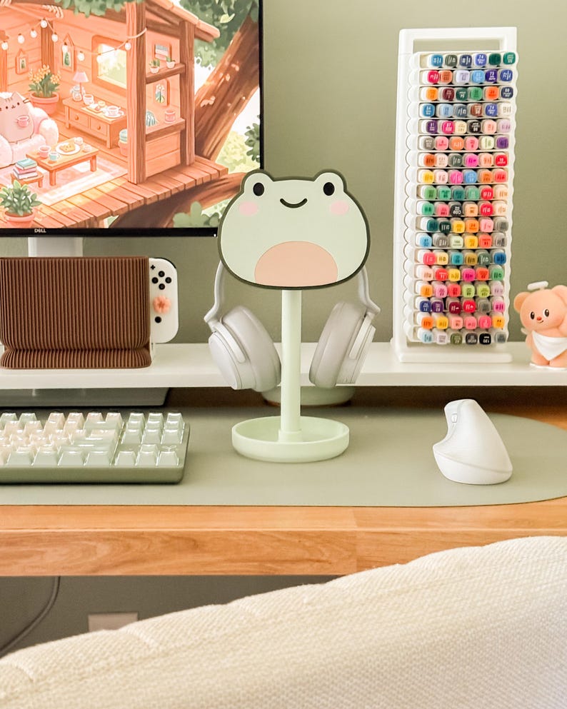 May include: A light green frog-shaped headphone stand holding white headphones. A white keyboard, mouse, and pen holder are on a desk. A white marker holder with colorful markers is in the background. A computer monitor displays a cartoon scene.