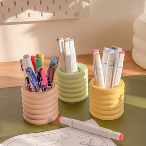 May include: Three colorful pen holders filled with markers and pens. The holders are in shades of tan, green, and yellow. A white marker with a red tip rests on a coloring page.