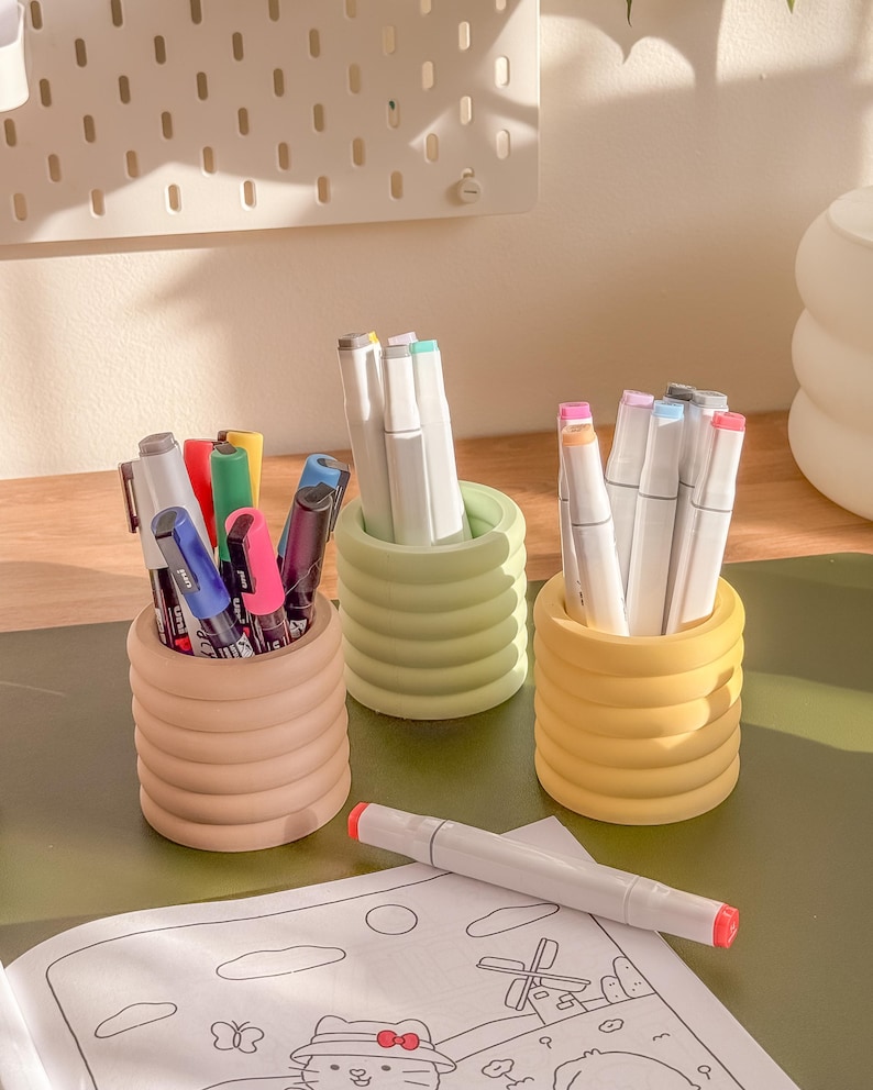 May include: Three colorful pen holders filled with markers and pens. The holders are in shades of tan, green, and yellow. A white marker with a red tip rests on a coloring page.