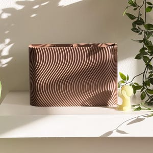 May include: A rectangular, brown planter with a wavy, textured design. The planter sits on a white shelf, with a small yellow candle and green leafy vines nearby. The shadows of the leaves are cast on the white wall.