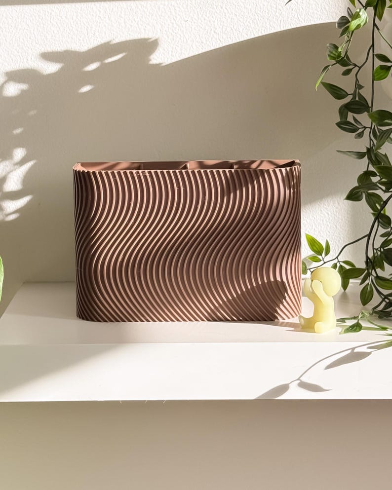 May include: A rectangular, brown planter with a wavy, textured design. The planter sits on a white shelf, with a small yellow candle and green leafy vines nearby. The shadows of the leaves are cast on the white wall.