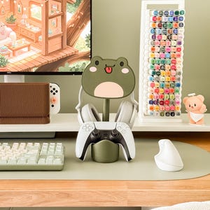 May include: A gaming setup featuring a white and black game controller in a green frog-shaped stand. White headphones rest on the stand. A white keyboard, mouse, and a marker holder with colorful markers are also visible.