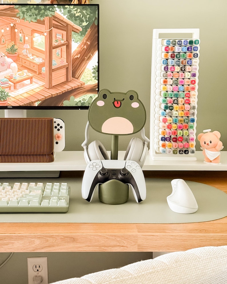 May include: A gaming setup featuring a white and black game controller in a green frog-shaped stand. White headphones rest on the stand. A white keyboard, mouse, and a marker holder with colorful markers are also visible.