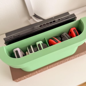 May include: A green and brown Nintendo Switch game console stand with slots for controllers. The stand has a brown base and a green top with five slots for controllers. There are four controllers in the slots, including two red, one white, and one black controller.