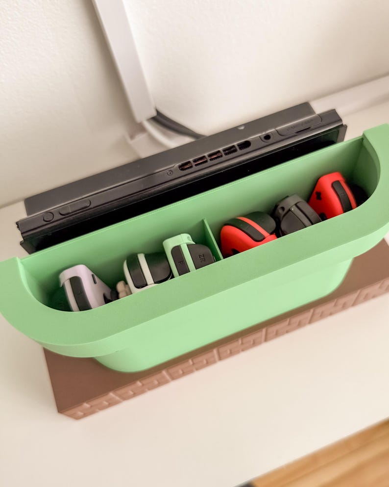 May include: A green and brown Nintendo Switch game console stand with slots for controllers. The stand has a brown base and a green top with five slots for controllers. There are four controllers in the slots, including two red, one white, and one black controller.