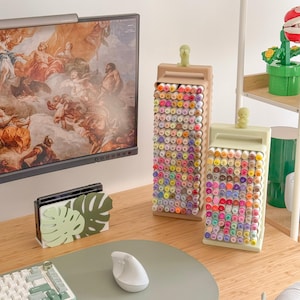 May include: Two rectangular marker holders filled with colorful markers sit on a desk. One holder is tan, the other is light green. Each has a small figurine on top. A computer monitor and a decorative monstera leaf stand are also visible.