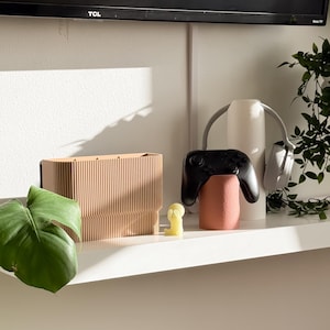 May include: A wooden rectangular box with vertical lines, a small yellow figurine, a black game controller, and a pink cylindrical object are on a white shelf. A pair of gray headphones and a green plant are also on the shelf. A black television is in the background.